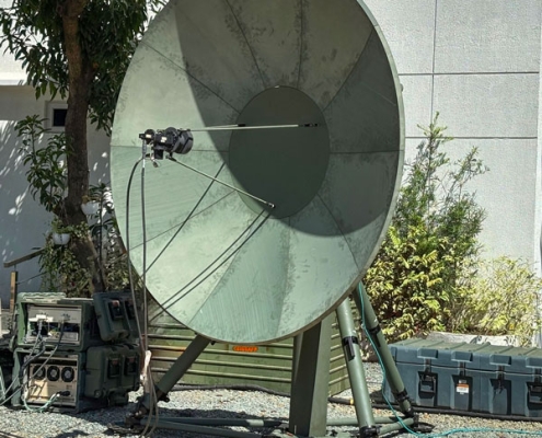 A close-up image of the MTTS Troposcatter communications system, featuring a large, green satellite dish mounted on a robust stand with supporting cables connected to electronic equipment, set outdoors in a gravel area with trees and a building wall in the background.