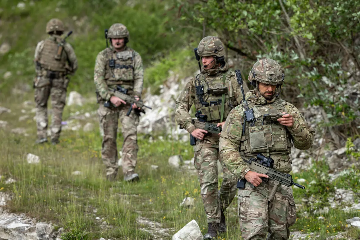 Four soldiers equipped with GENSS, operating in the field, with a foreground soldier using tactical communications equipment.